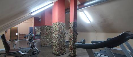 Un gimnasio con equipamiento de ejercicio, incluyendo cintas de correr y bicicletas estáticas, bajo un techo inclinado con iluminación fluorescente.