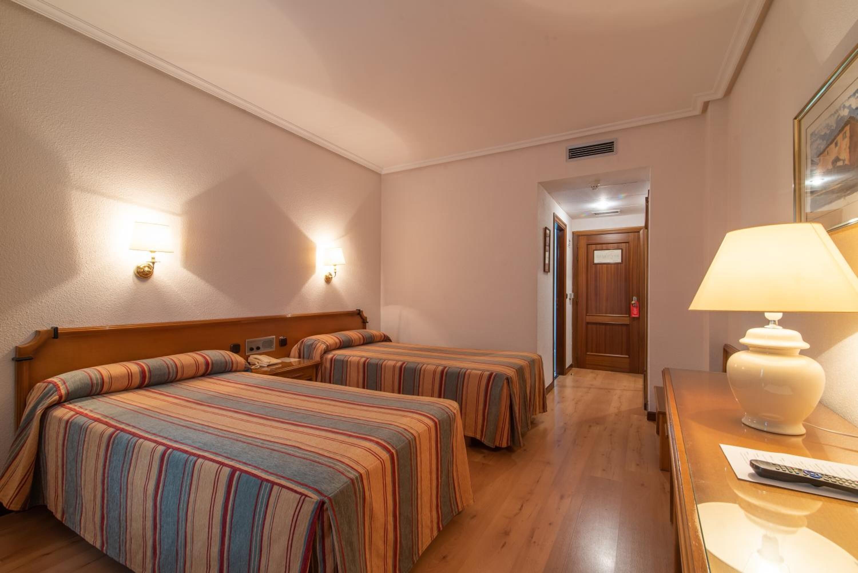 Una habitación de hotel con dos camas, un escritorio con lámpara y una puerta que da a otra habitación.