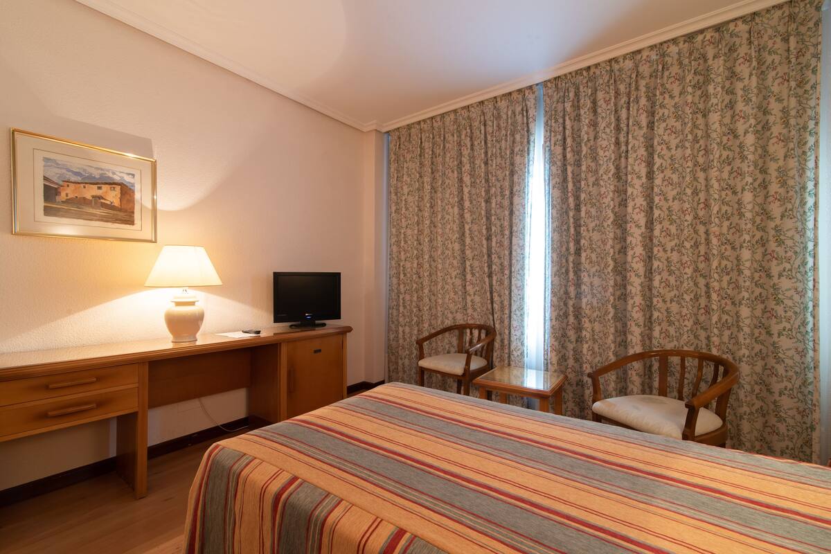 A hotel room with a bed, a desk with a lamp, a television, two chairs, and a window with floral curtains.
