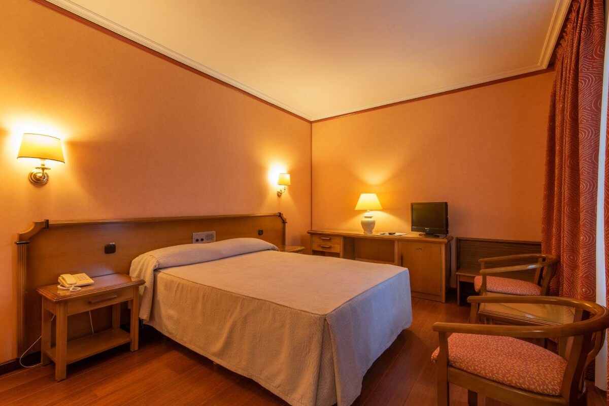 Una habitación de hotel con una cama, mesitas de noche, un escritorio con lámpara, una silla y un televisor.