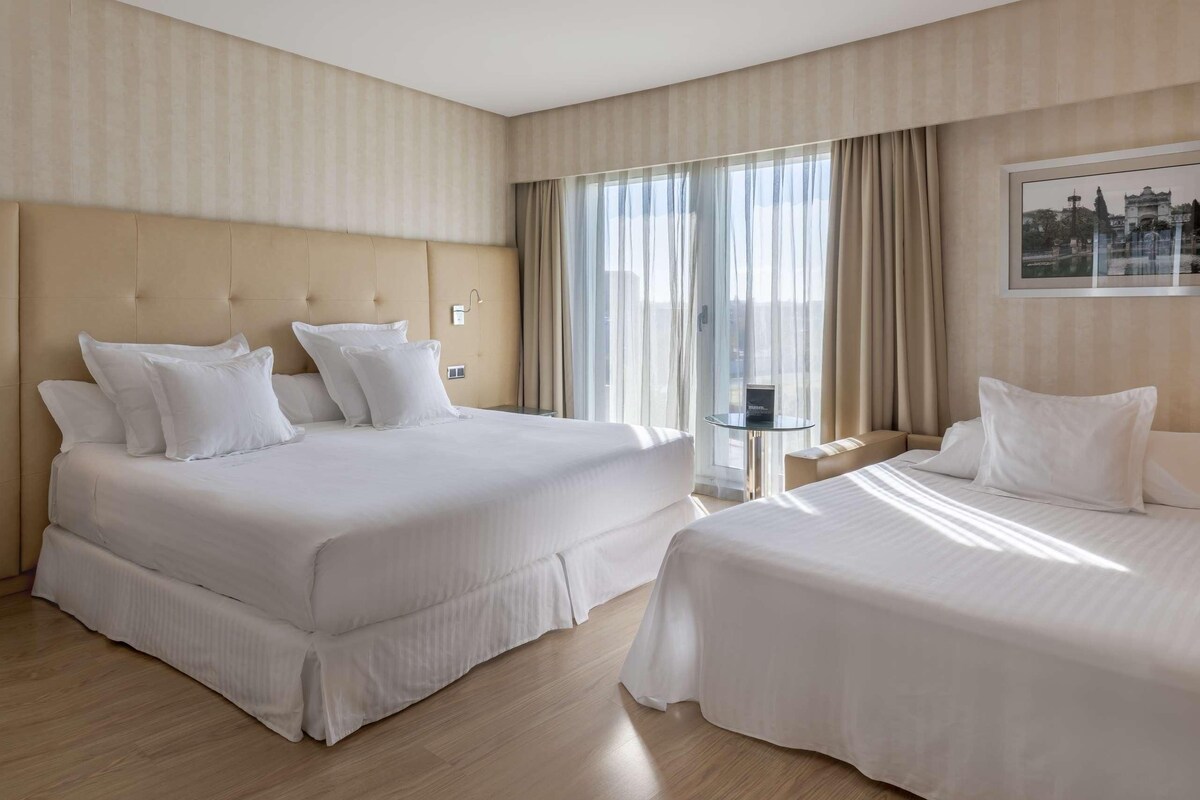 A hotel room with two beds, a large window with curtains, a small table, and a framed picture on the wall.