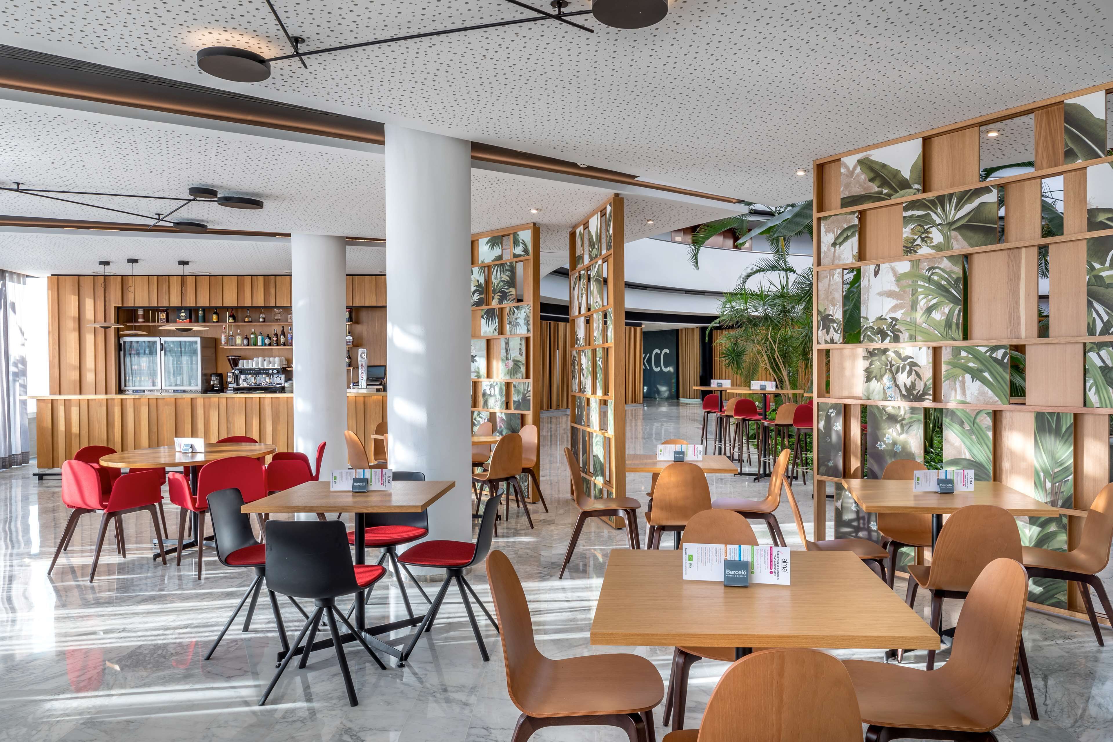 A modern indoor space with wooden tables and chairs, a bar area, and large windows with tropical plant views.