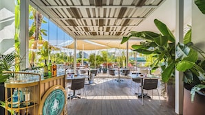 Un patio al aire libre con mesas y sillas, una zona de bar con botellas y plantas en macetas grandes.