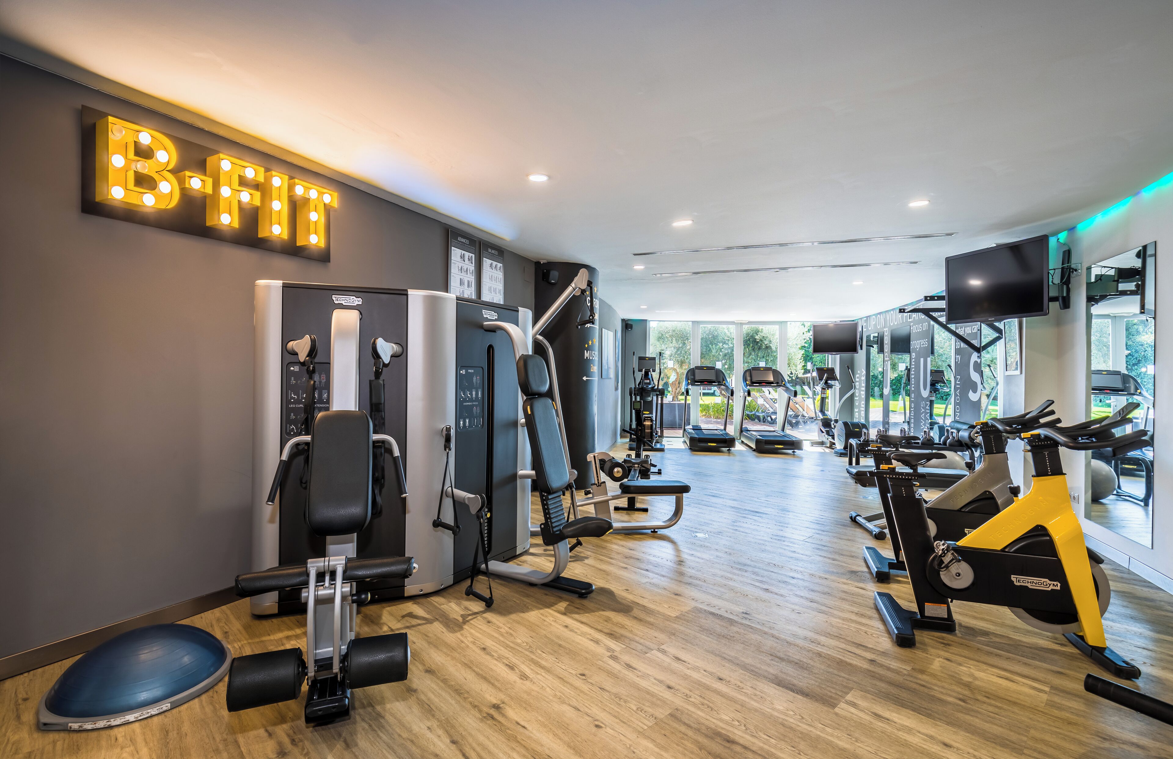 Une salle de sport équipée de divers appareils d’exercice, une grande enseigne lumineuse sur laquelle on peut lire « B-FIT », et une vue sur l’extérieur grâce à de grandes fenêtres.