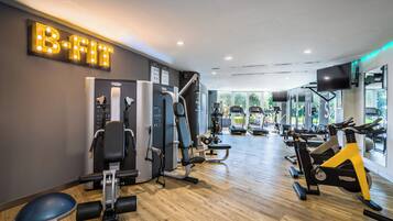 Une salle de sport équipée de divers appareils d’exercice, une grande enseigne lumineuse sur laquelle on peut lire « B-FIT », et une vue sur l’extérieur grâce à de grandes fenêtres.
