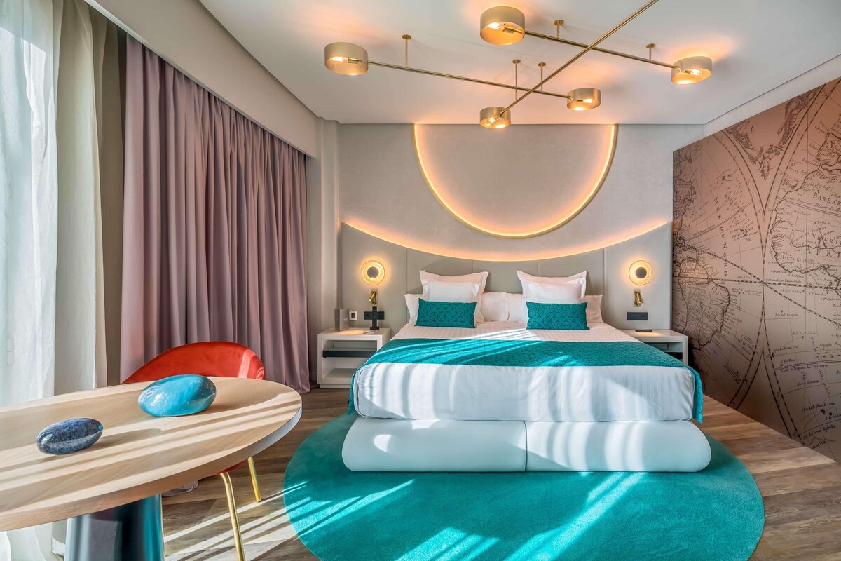 A modern hotel room with a large bed, a desk, and a chair. There are wall-mounted lamps and a map on the wall.