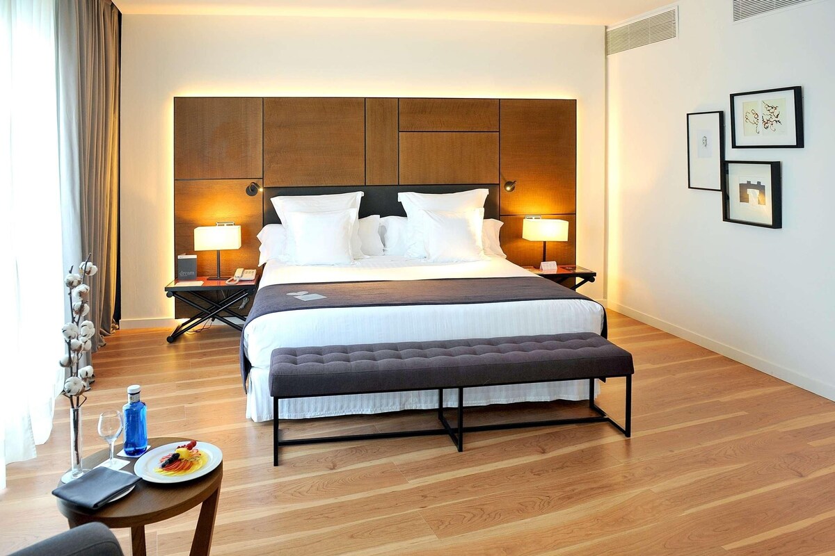 A hotel room with a large bed, bedside tables with lamps, a bench, a small table with a plate of food, and a vase of flowers.
