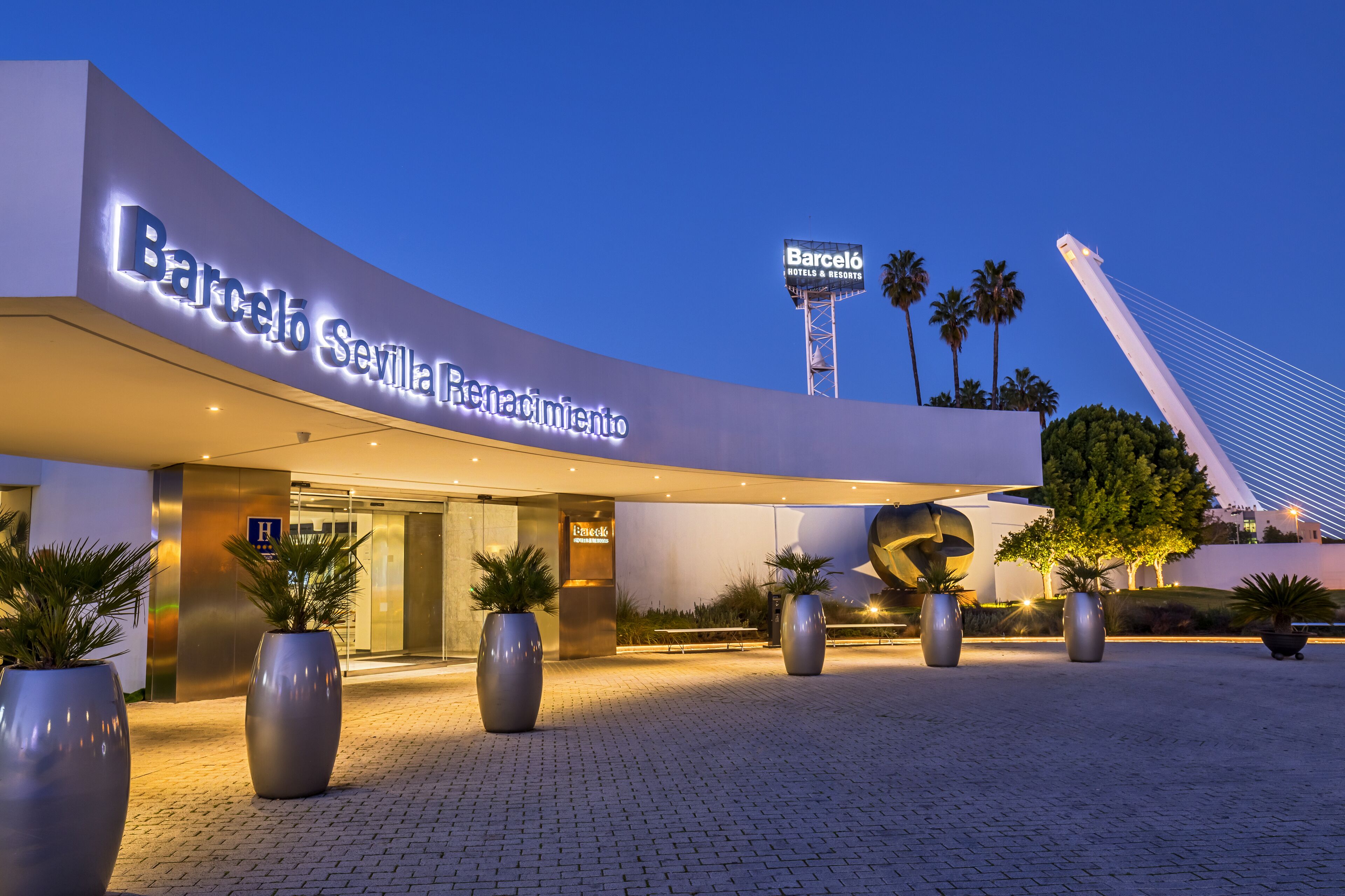 The exterior of Barceló Sevilla Renovación hotel with a modern design, large potted plants, and a prominent tower in the background.