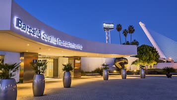 The exterior of Barceló Sevilla Renovación hotel with a modern design, large potted plants, and a prominent tower in the background.