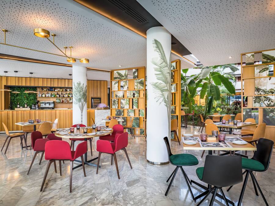 Un restaurante moderno con suelo de mármol, sillas rojas y verdes, y una zona de bar con armarios de madera y plantas.