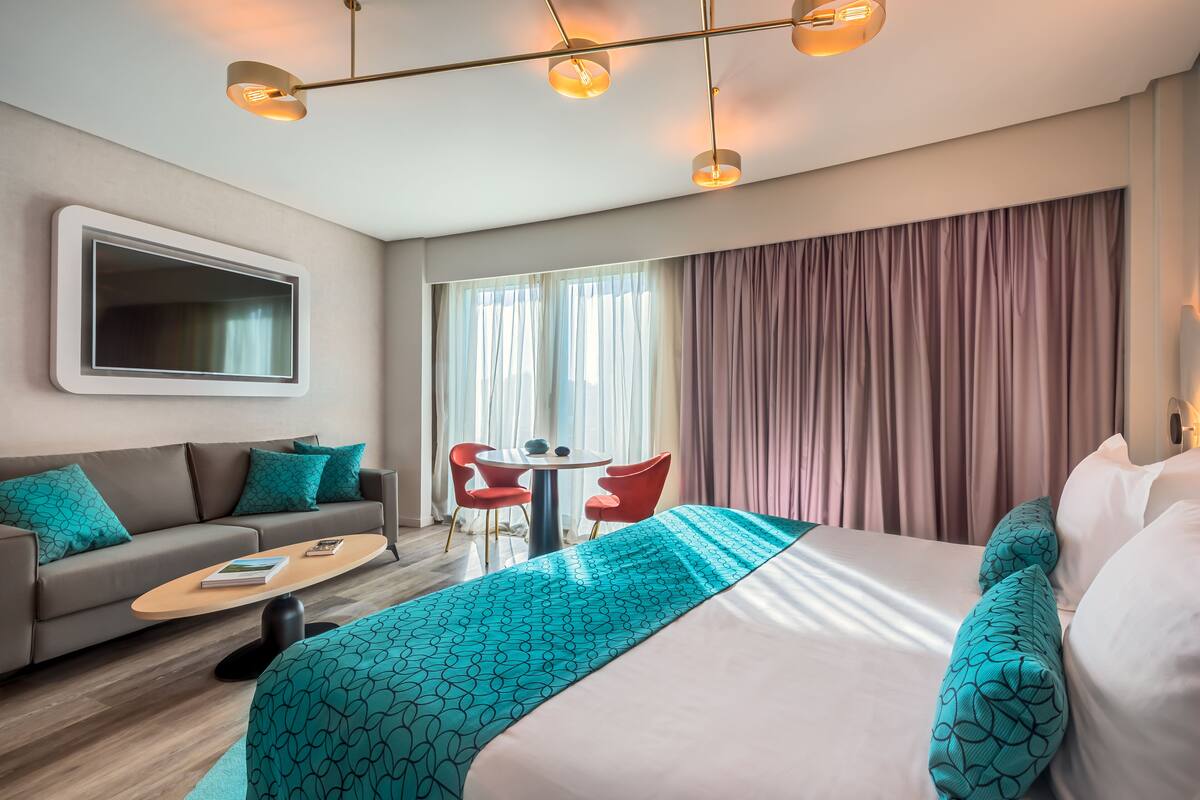 A modern hotel room with a large bed, a sofa, a coffee table, and a desk with chairs.