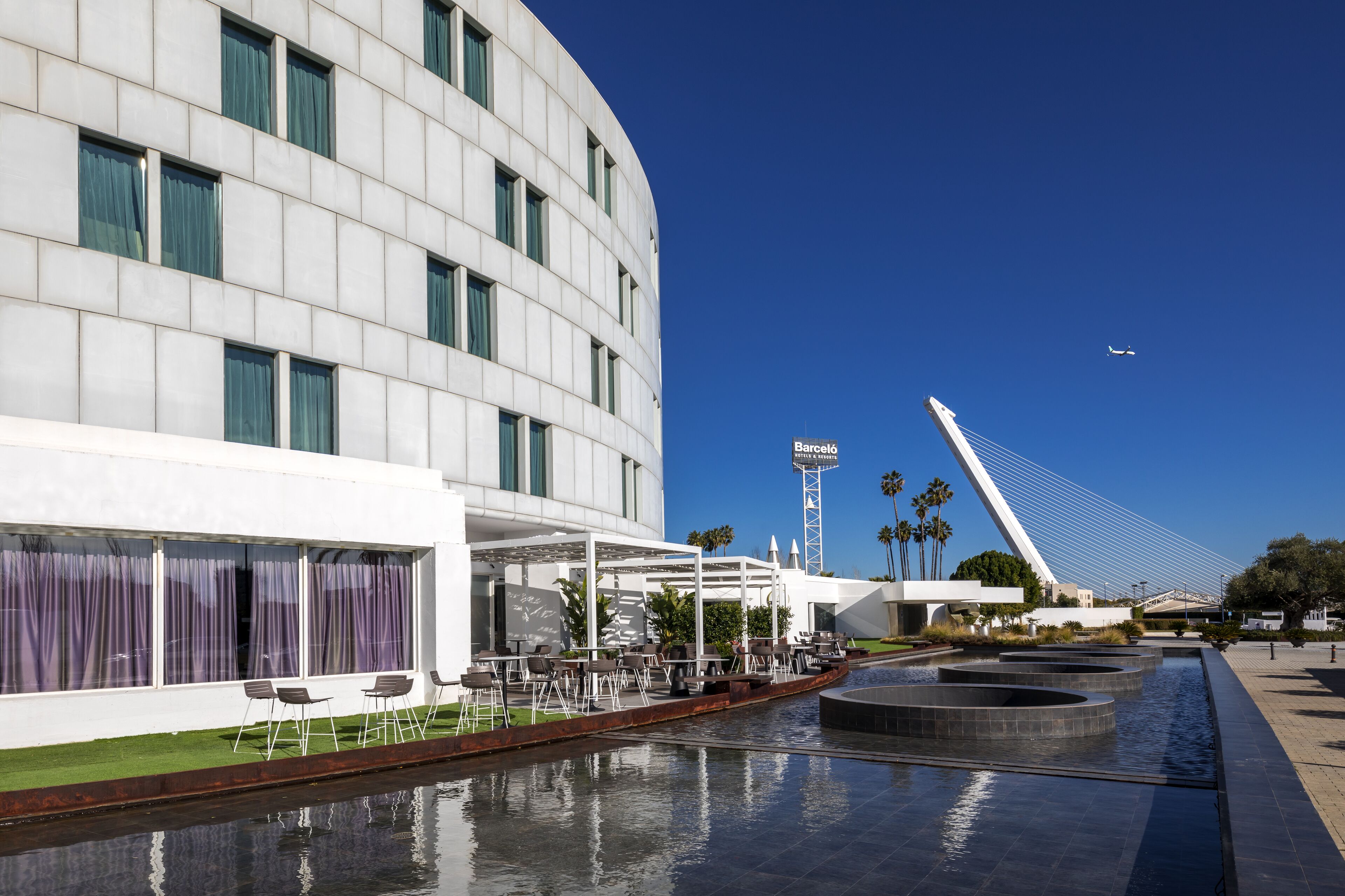 A modern hotel with a curved facade, outdoor seating area, and a water feature.