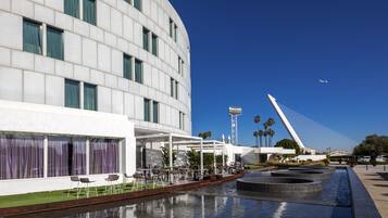 A modern hotel with a curved facade, outdoor seating area, and a water feature.
