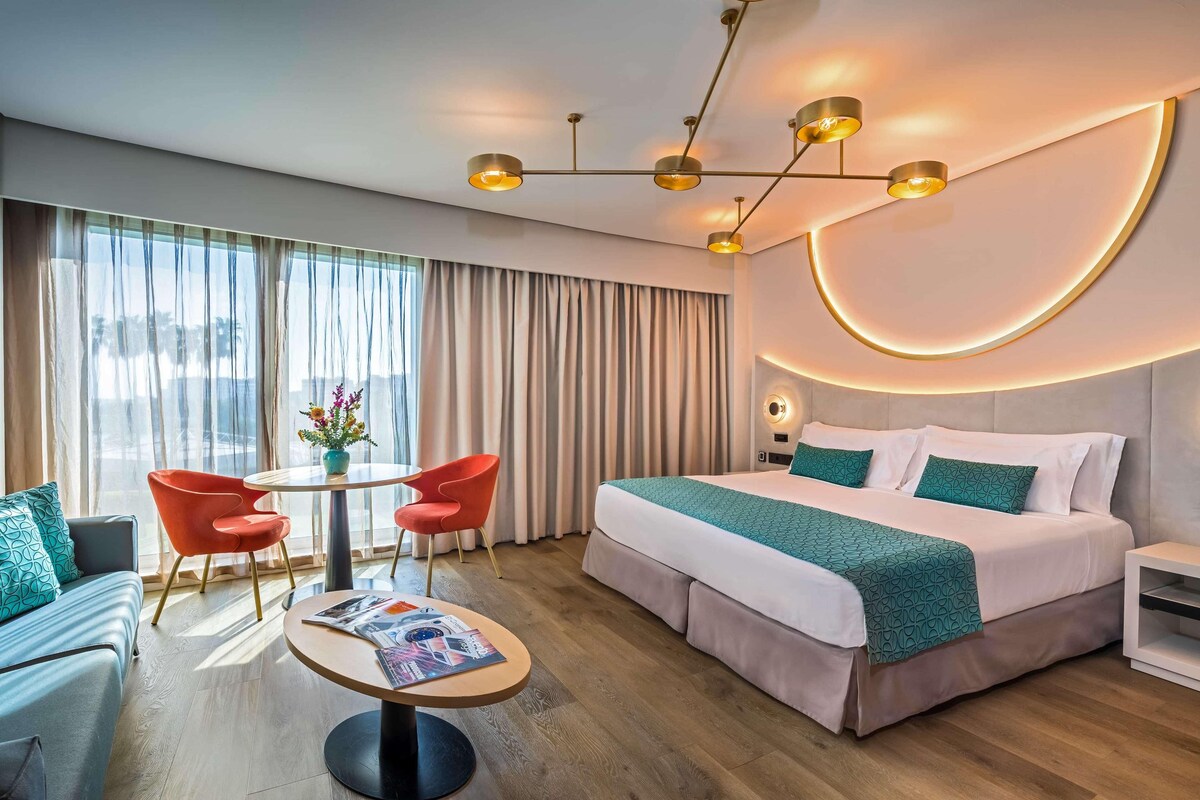A modern hotel room with a large bed, a dining table with chairs, and a sofa.