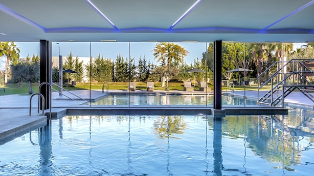 Una piscina interior con paredes de vidrio transparente, un suelo de baldosas y vistas a un jardín ajardinado con árboles y césped.