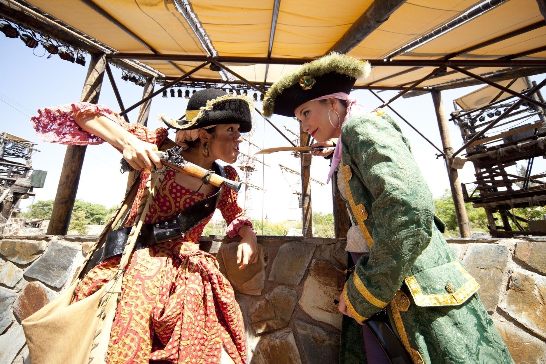 Two people in historical costumes under a wooden structure with stone walls.
