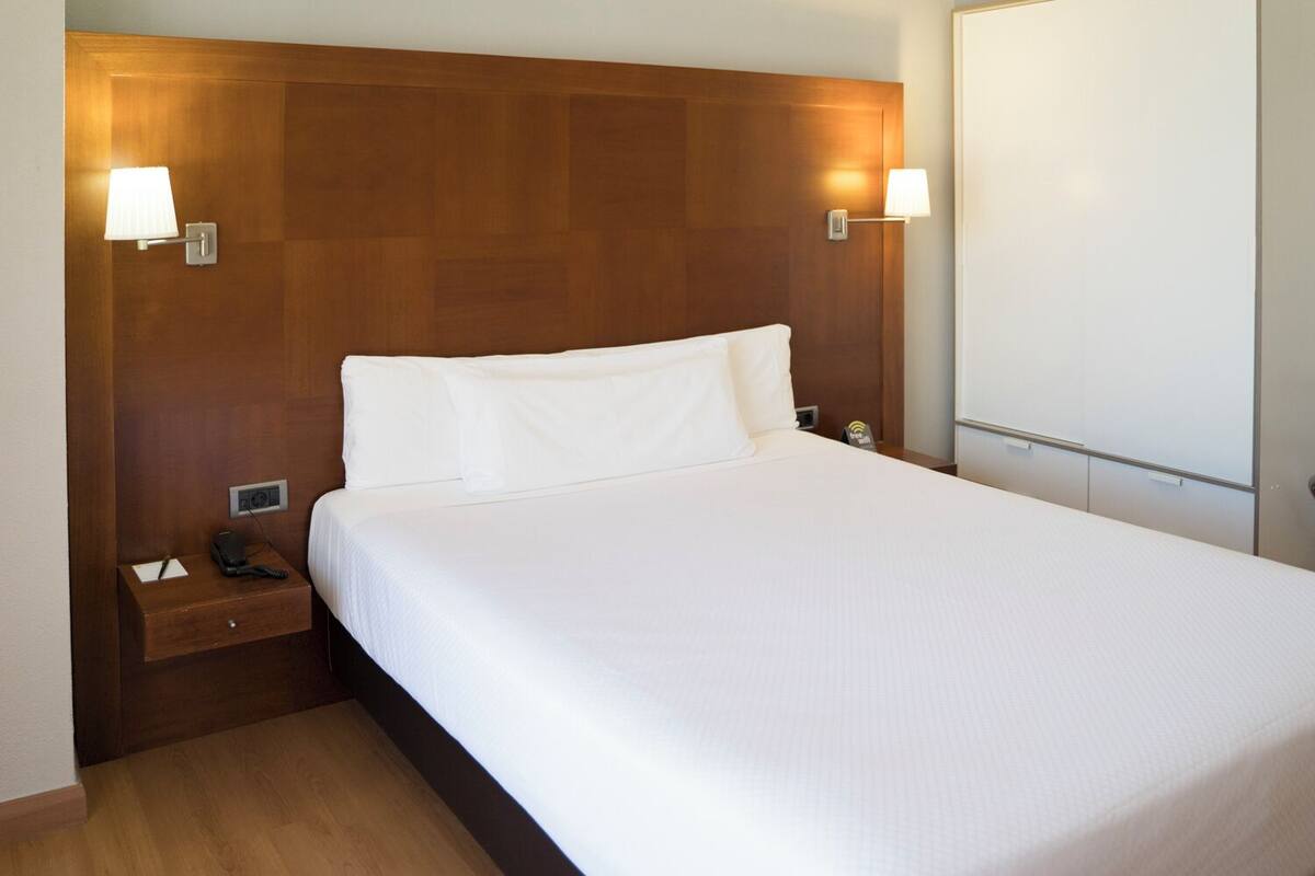 A neatly made bed with white linens, a wooden headboard, bedside lamps, and a nightstand with a phone and a tissue box.