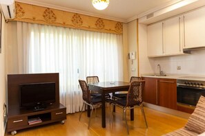 Un espacio habitable compacto con una TV sobre un mueble de madera, una mesa de comedor con cuatro sillas, una zona de cocina con armarios y una estufa, y un ventanal con cortinas.