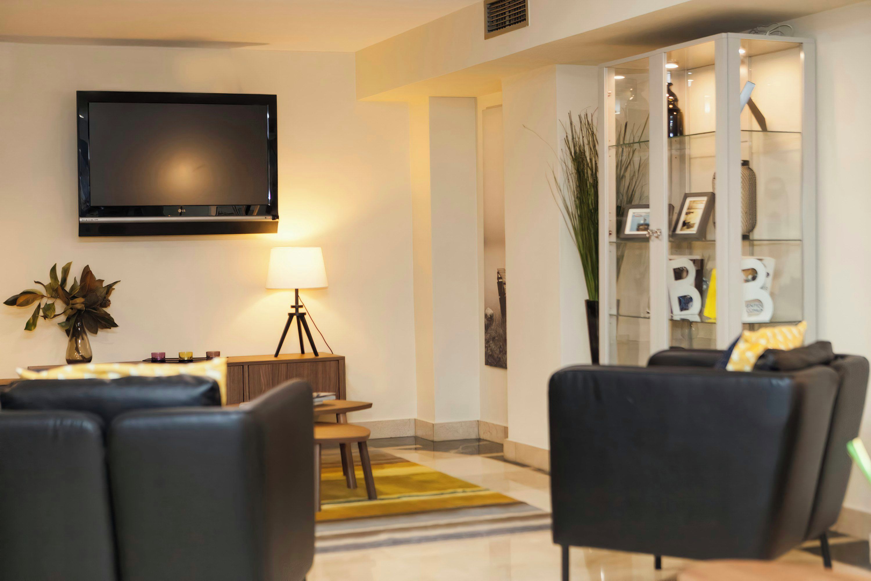 A modern living room with a flat-screen TV, a lamp, a small table with candles, and a glass cabinet with decorative items.