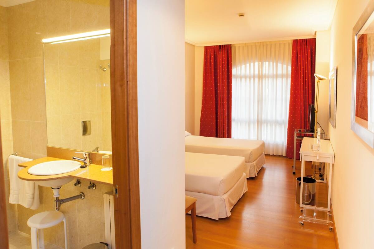 A hotel room with a bathroom, two beds, a bedside table, a lamp, and a window with curtains.