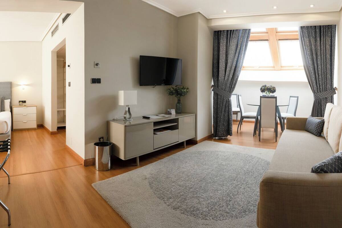 A modern living room with a sofa, a TV, a dining table with chairs, and a small kitchen area.