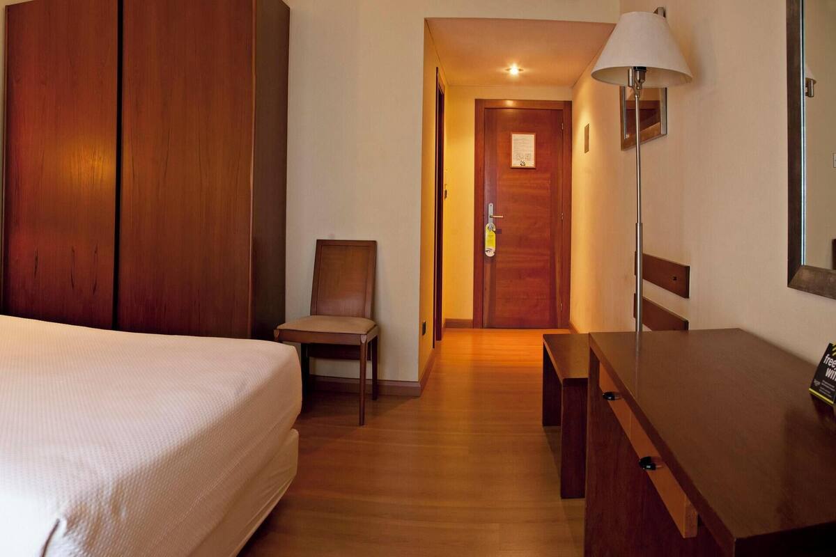 A hotel room with a large bed, a wooden wardrobe, a desk, and a chair. There are two wall-mounted lamps and a telephone on the nightstand.