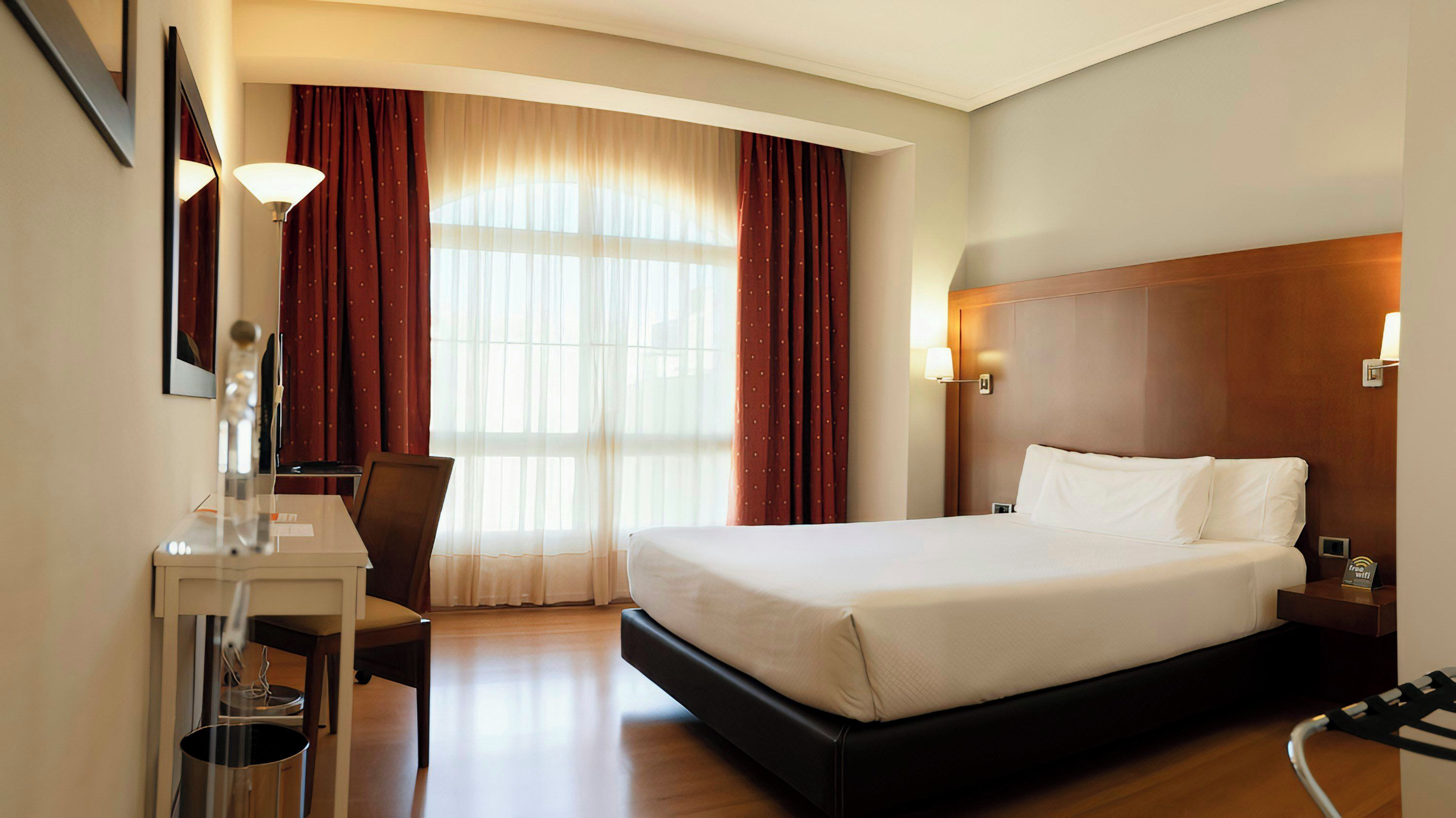 Quarto de hotel com cama, escrivaninha, cadeira, luminária e janela com cortinas.