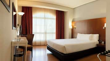 Quarto de hotel com cama, escrivaninha, cadeira, luminária e janela com cortinas.