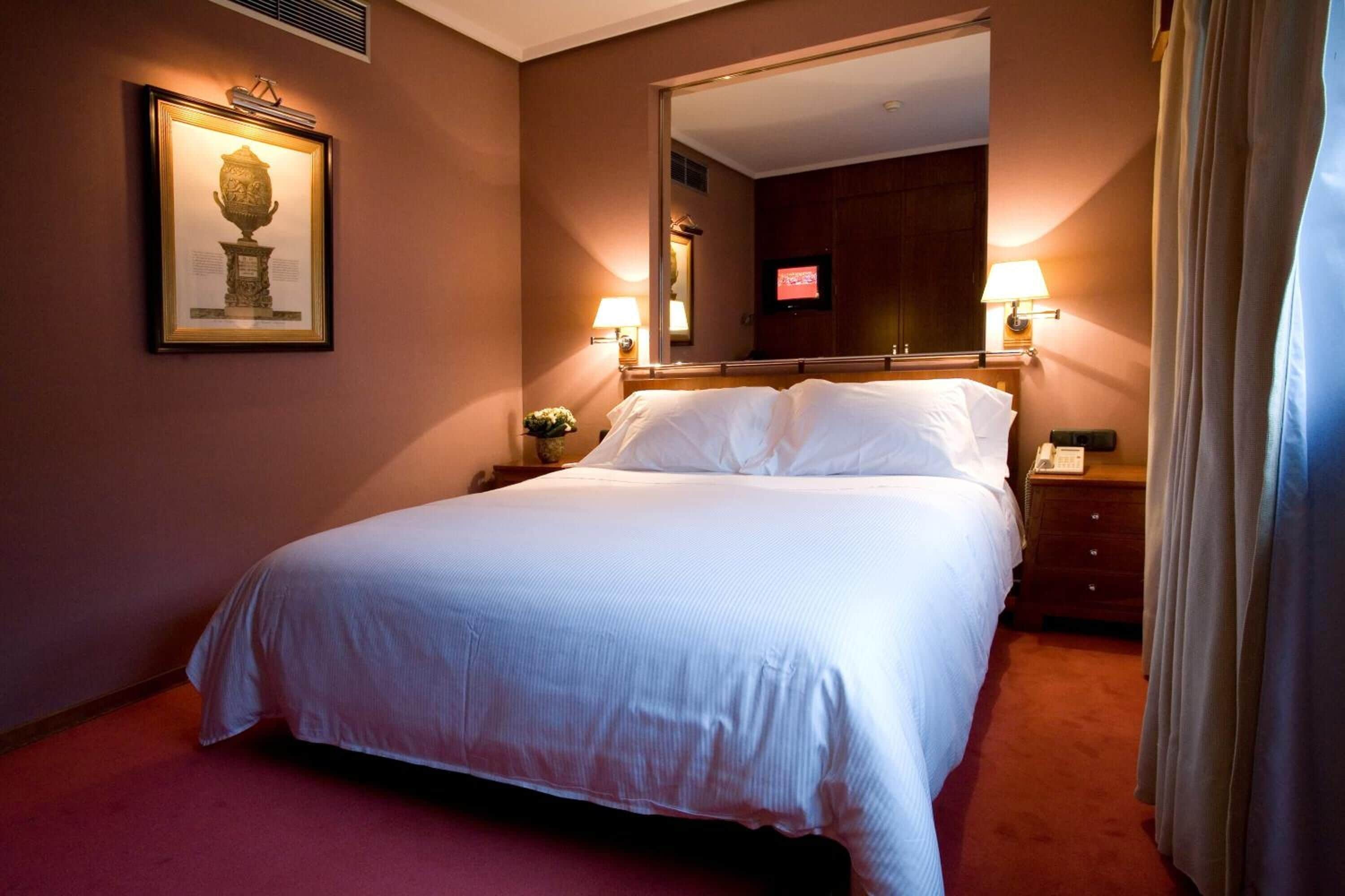 Habitació d'hotel amb un llit gran, una nit falda amb una làmpada, un quadre amb marc a la paret i un mirall que reflecteix l'habitació.