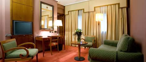 A hotel room with a television, a desk, a chair, a sofa, and a lamp.