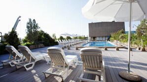 A poolside area with lounge chairs and umbrellas, a pool, and a modern building in the background. - Hotel Palafox (Zaragoza)