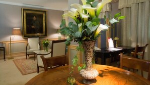 A dining area with a round wooden table, a vase with flowers, a framed portrait on the wall, and a lamp on a side table. - Hotel Palafox (Zaragoza)