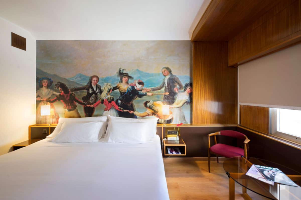 Una habitación de hotel con una cama grande, una mesita de noche de madera, una silla roja, una mesa de vidrio y un cuadro en la pared.