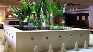 A lobby with a modern geometric light fixture, a central planter with green plants, and a seating area with tables and chairs. - Hotel Goya (Zaragoza)
