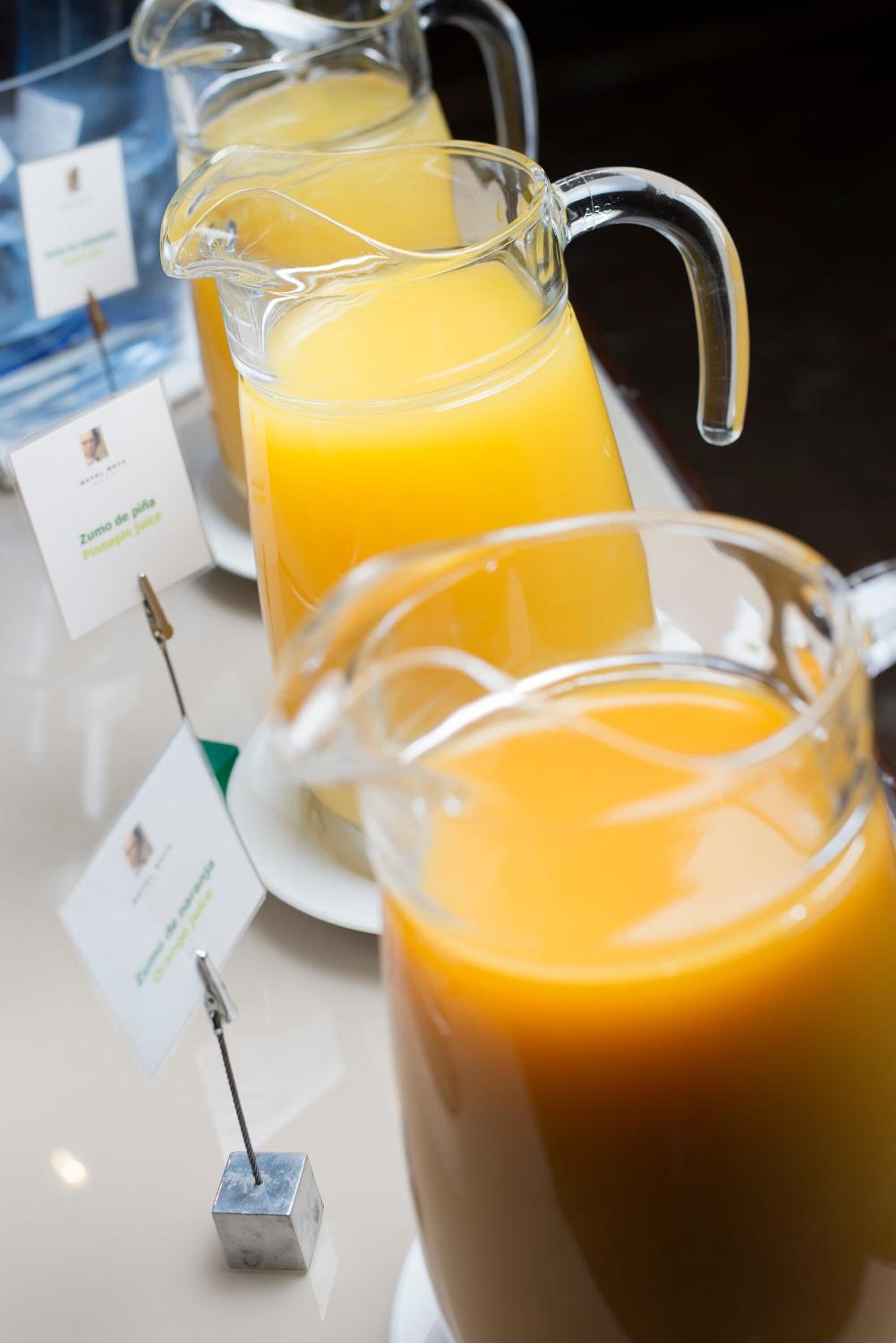 Dos jarras de jugo de naranja sobre una mesa con letreros que indican el tipo de jugo.