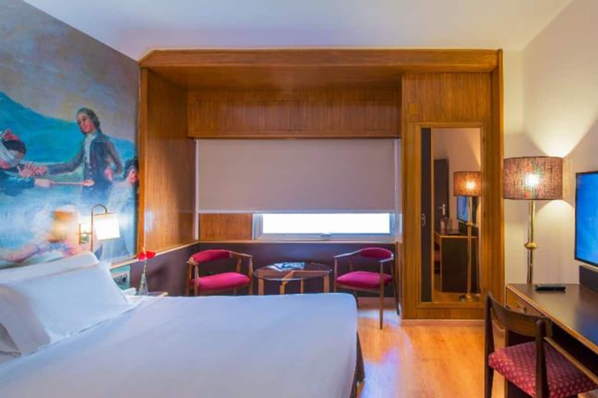 Habitación de hotel con cama, escritorio con televisor, dos sillas, ventana y un cuadro en la pared.
