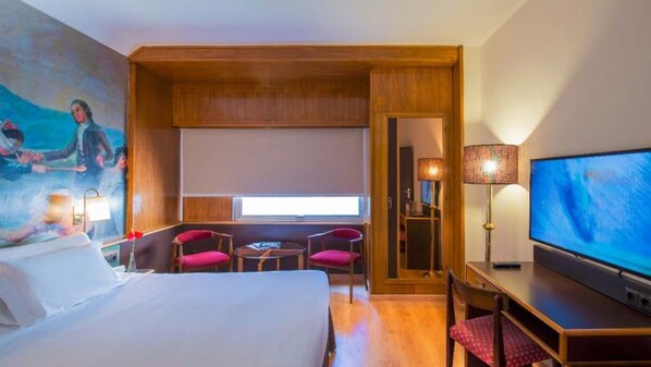 A hotel room with a bed, a desk with a TV, two chairs, a window, and a painting on the wall. - Hotel Goya (Zaragoza)