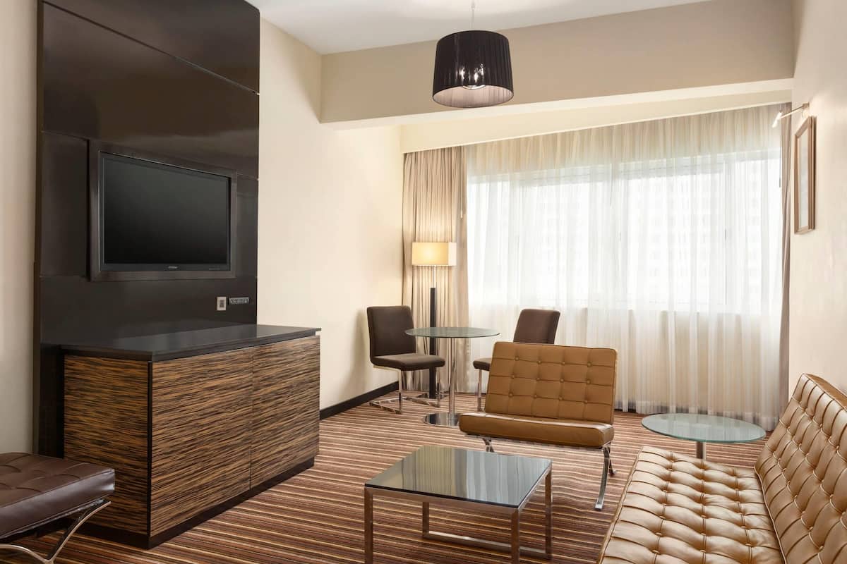 A hotel room with a flat-screen TV, a sofa, a glass coffee table, a small round table, and a lamp.