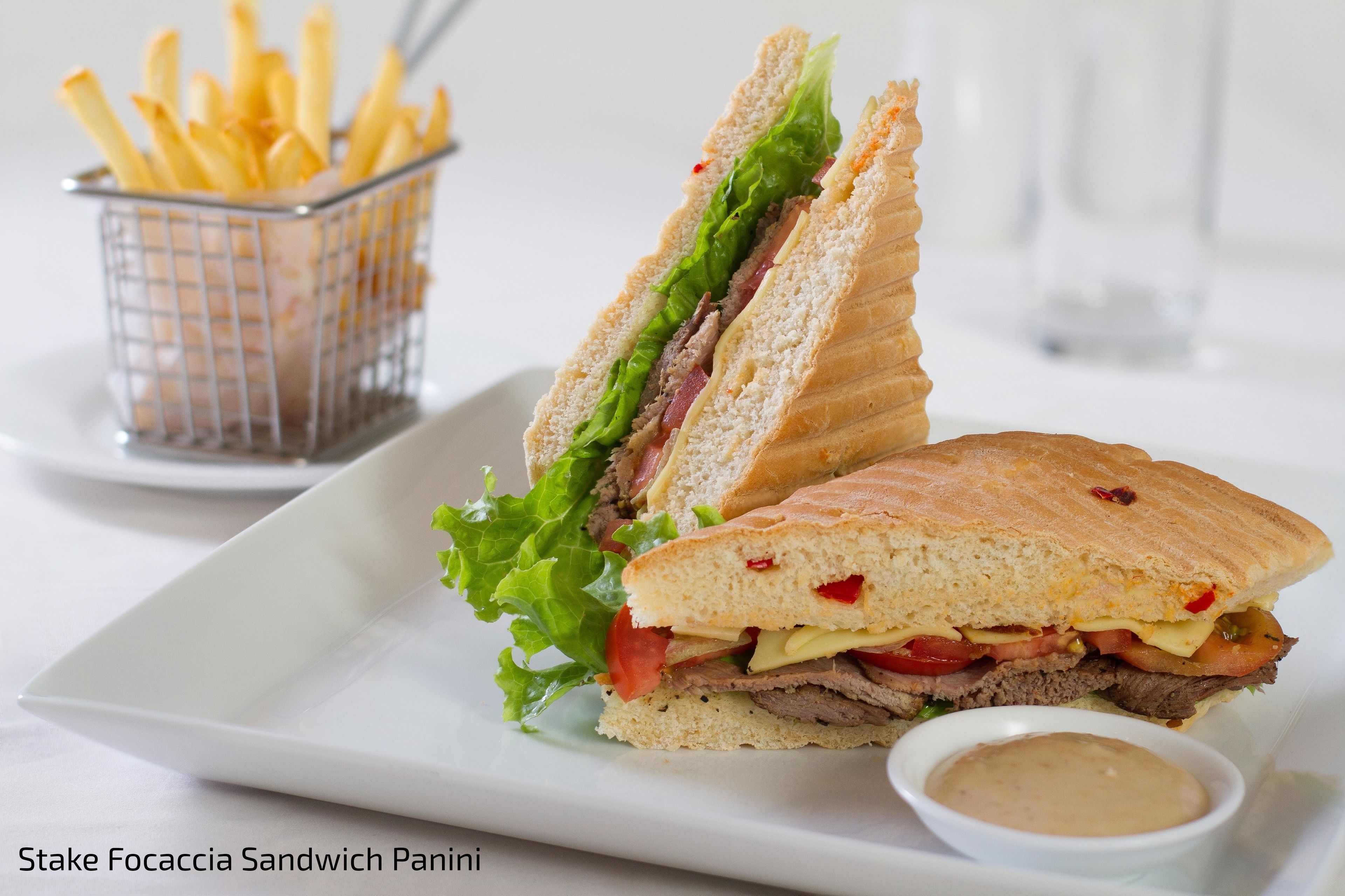A sandwich with lettuce, tomato, and meat, served on a white plate with a side of fries and a small bowl of sauce.