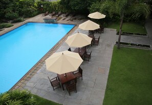 Uma piscina com um deck ladrilhado, rodeada por área de estar ao ar livre com guarda-sóis e um gramado bem cuidado.