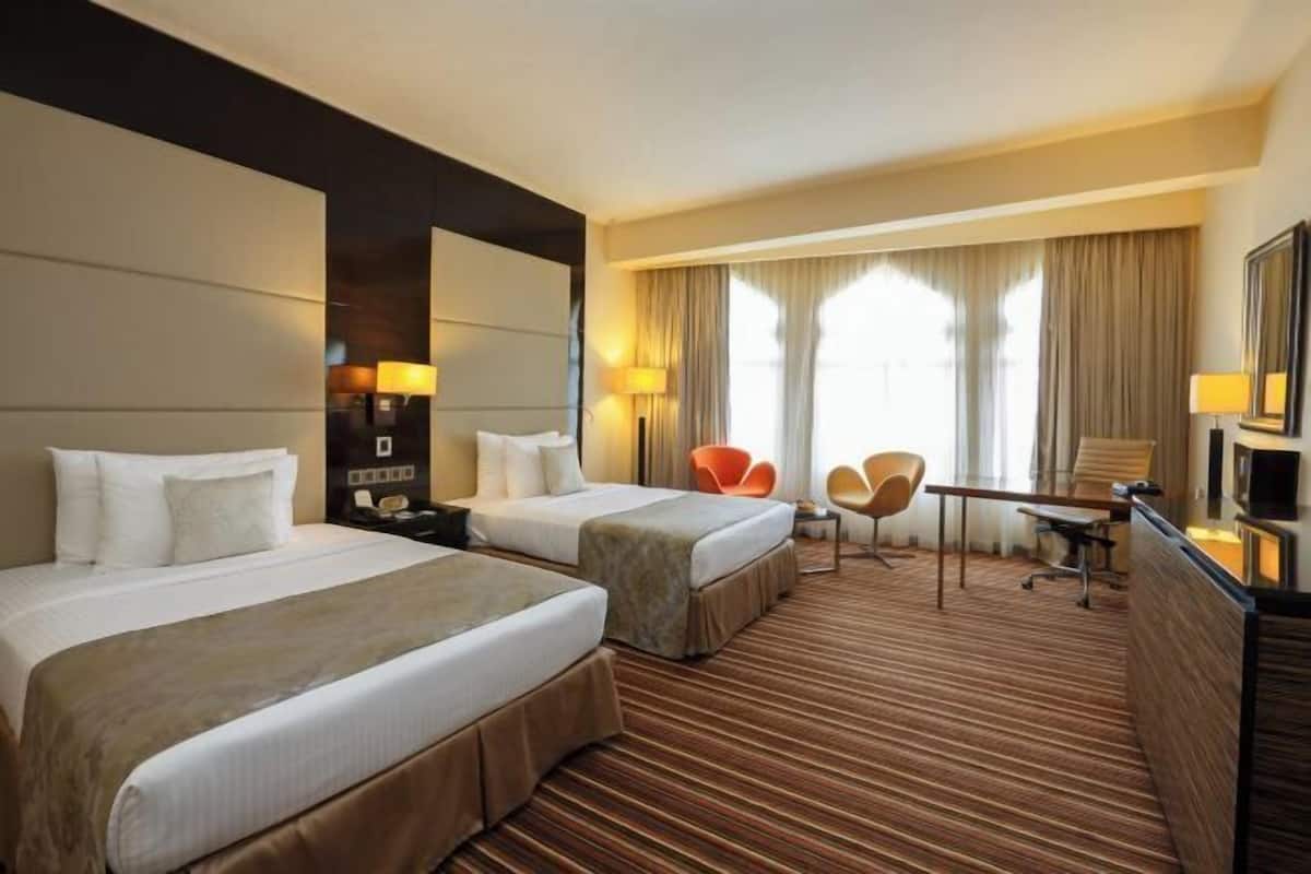A hotel room with two beds, a desk, a chair, a TV, and a large window with curtains.