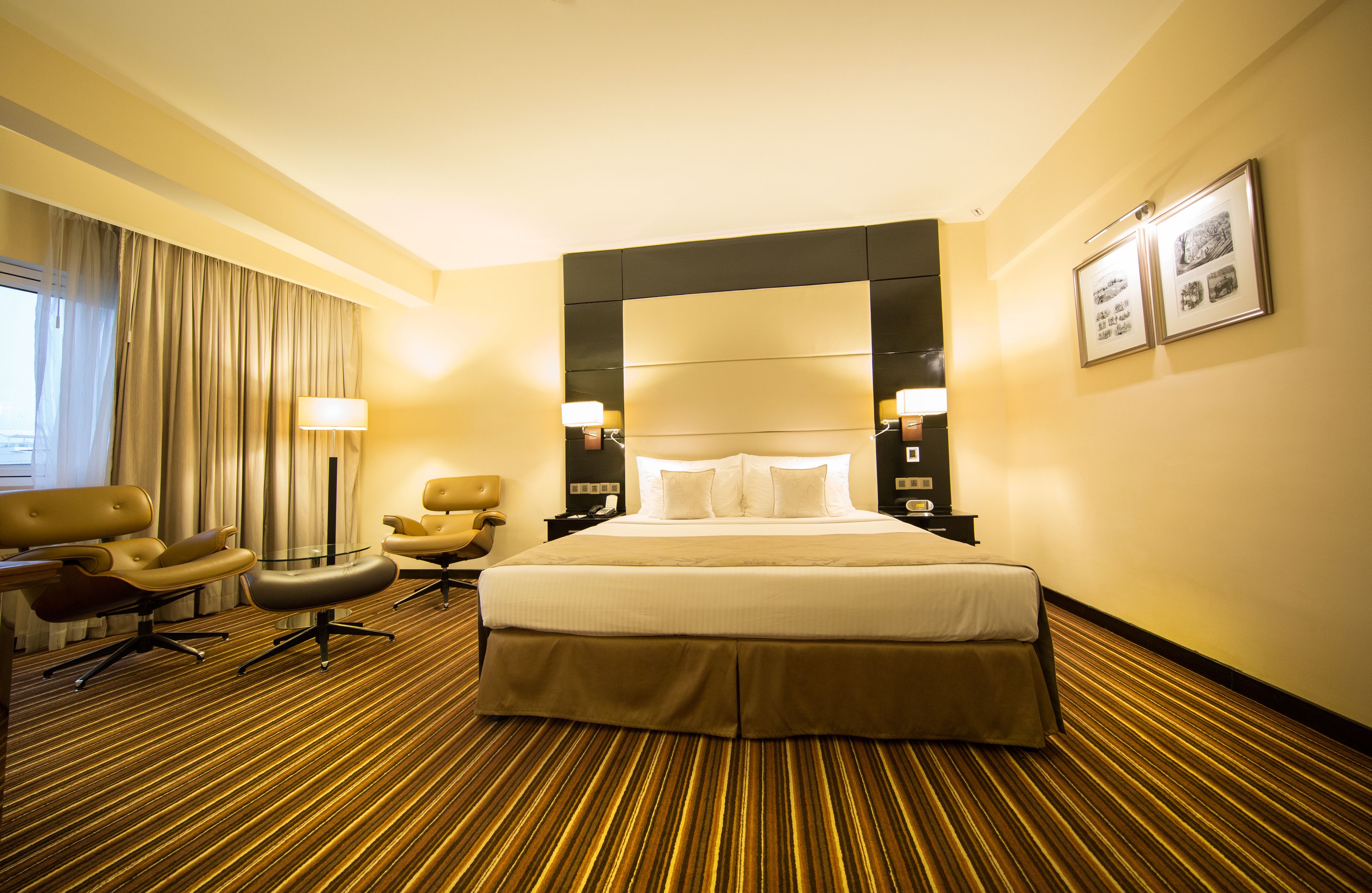 Une chambre d’hôtel avec un grand lit, deux fauteuils, une petite table et une fenêtre avec des rideaux.