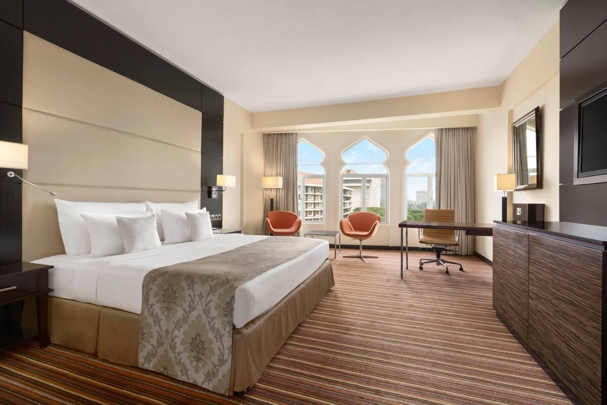 A hotel room with a large bed, a desk, two chairs, a TV, and a view of the city.