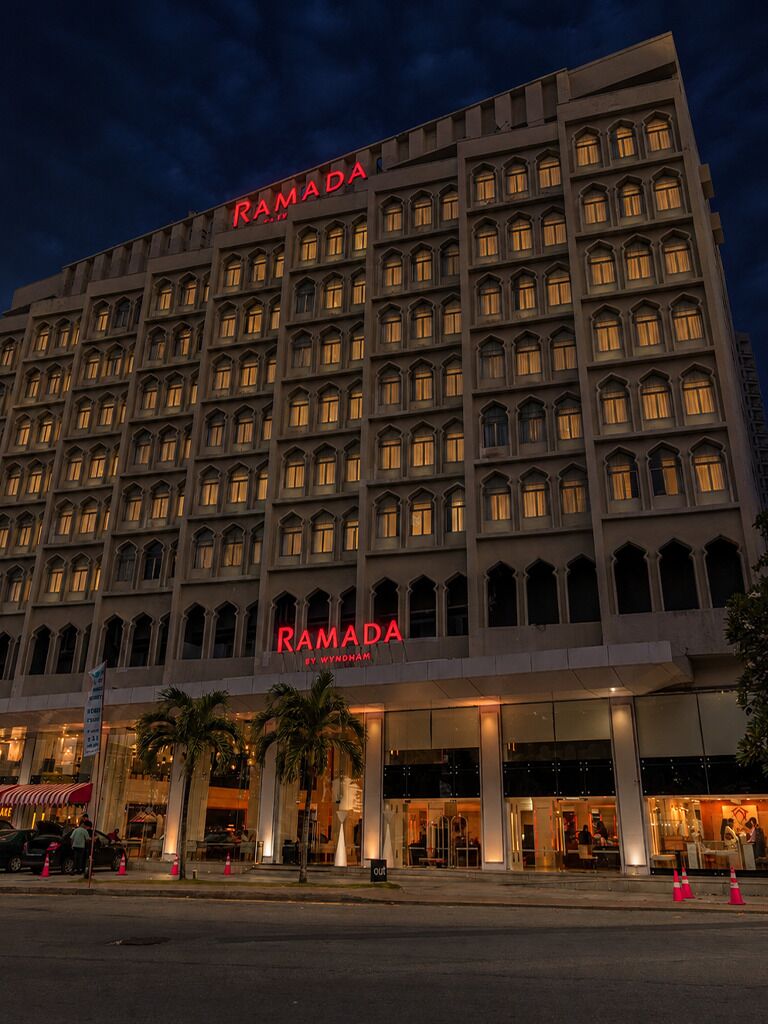 Ein mehrgeschossiges Hotel mit einem gut sichtbaren 'RAMADA'-Schild an der Fassade, mit einem symmetrischen architektonischen Design, zahlreichen Fenstern und einem flachen Dach.