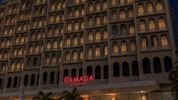 Un hotel de varios pisos con un llamativo letrero de 'RAMADA' en la fachada, que presenta un diseño arquitectónico simétrico con numerosas ventanas y un techo plano.