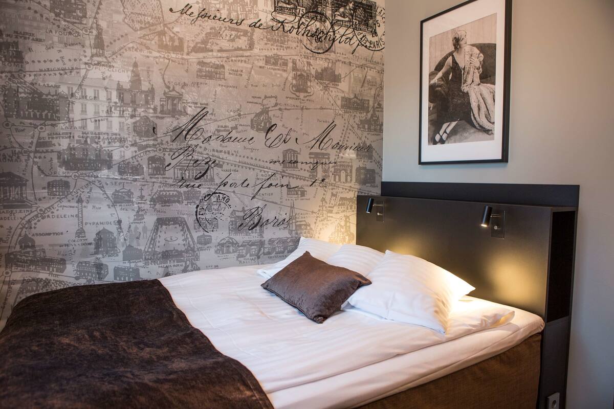A neatly made bed with a brown pillow and a blanket, a headboard with a light fixture, and a wall with a map and a framed picture.