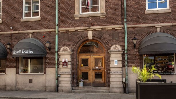 A brick building with a double door entrance, a sign reading "Hotel Borås," and a planter with plants on the sidewalk. - Hotell Boras, BW Signature Collection (Borås)