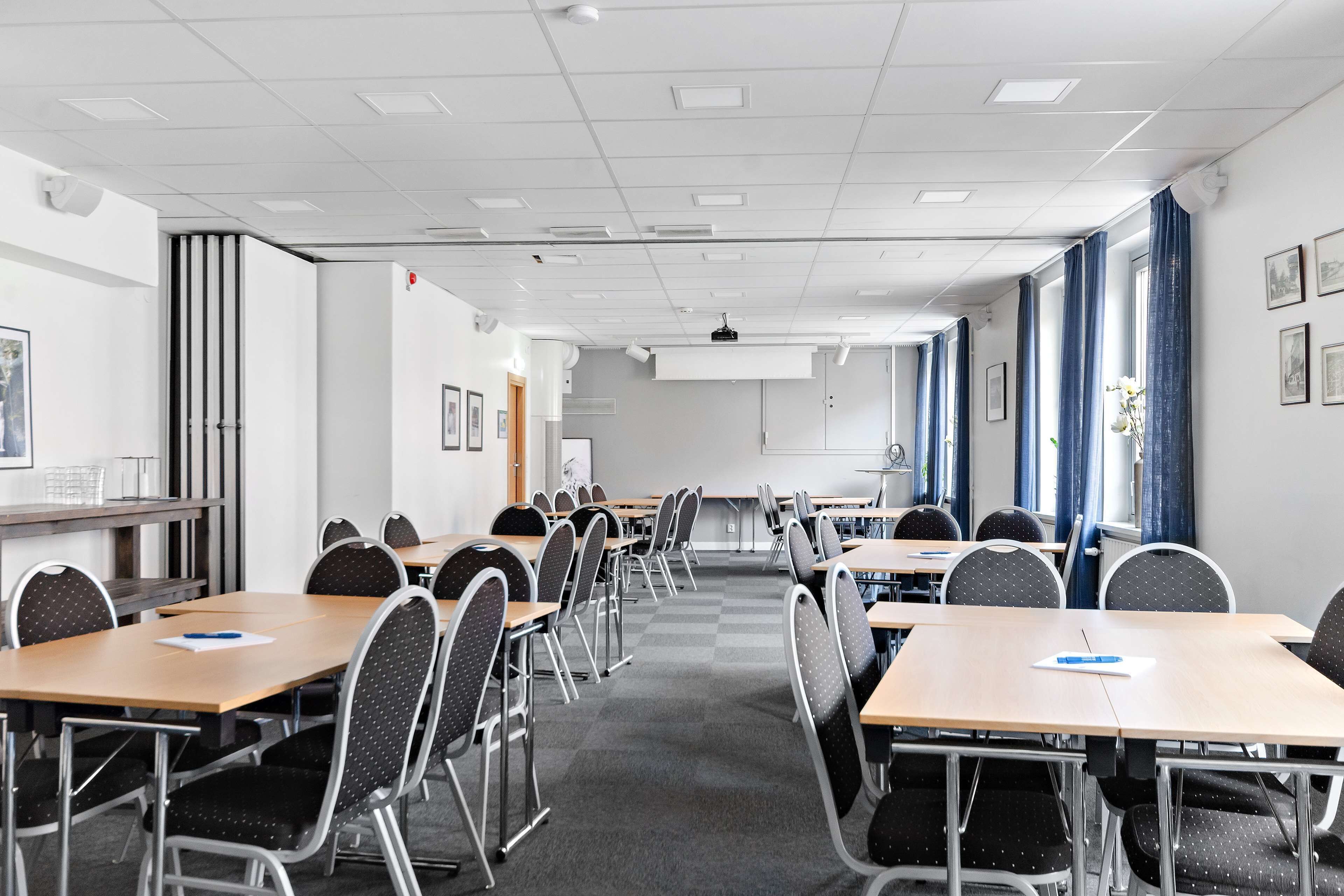A conference room with multiple tables and chairs, white walls, and a projector screen.