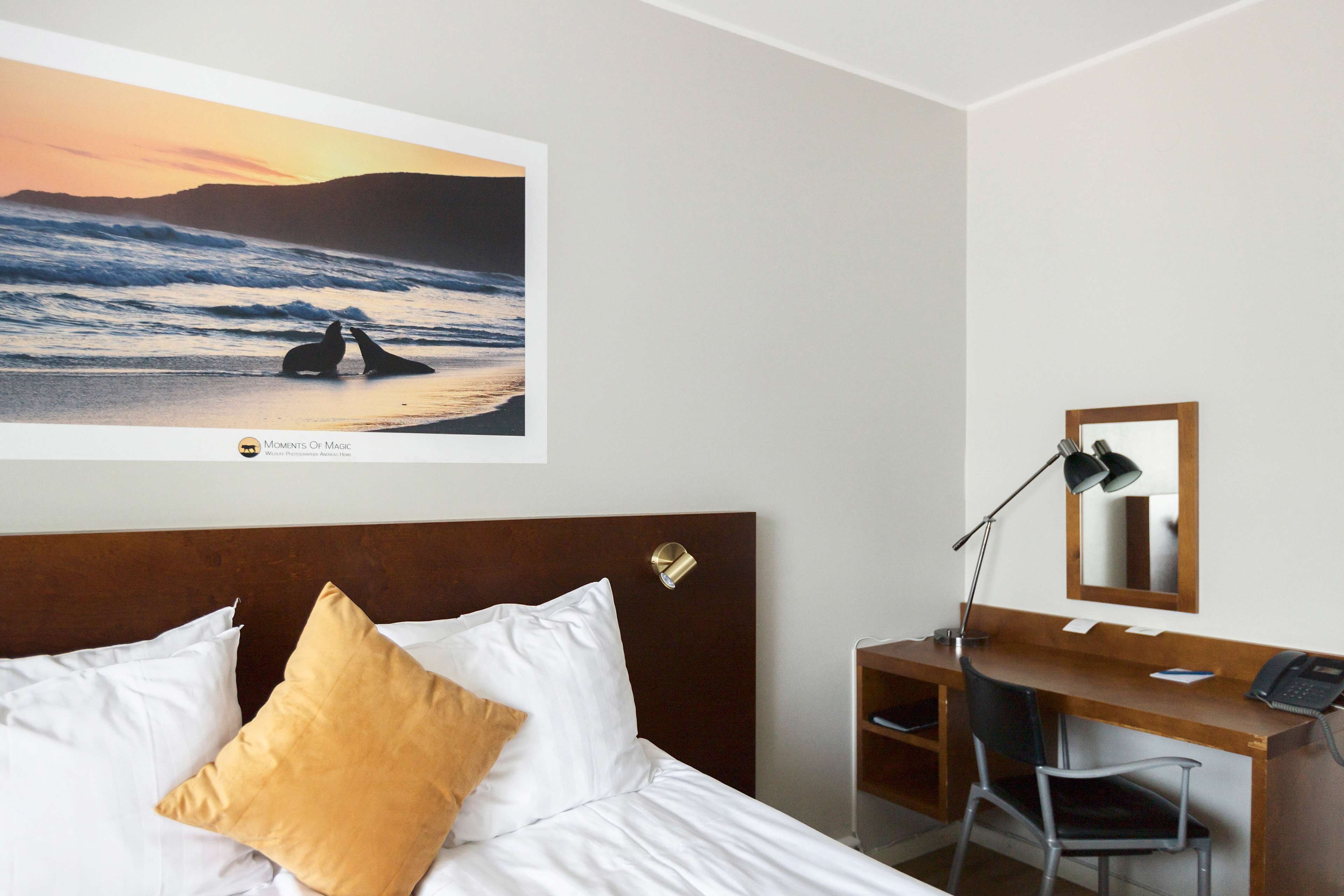 A hotel room with a bed, a desk, a chair, a desk lamp, a phone, and a framed picture of a beach scene.