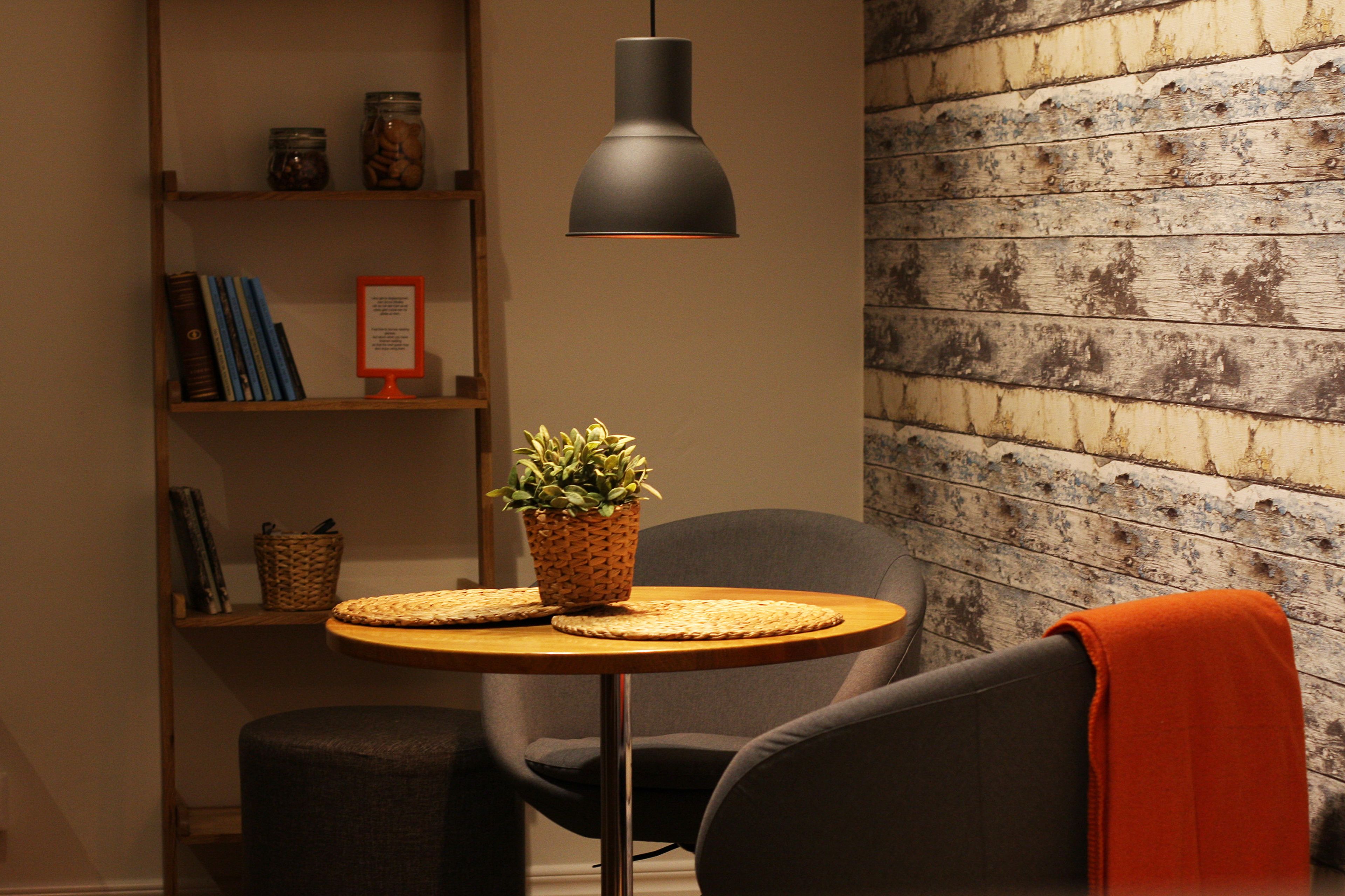 A cozy room with a round table, two chairs, a bookshelf, and a wall with a rustic wooden texture.